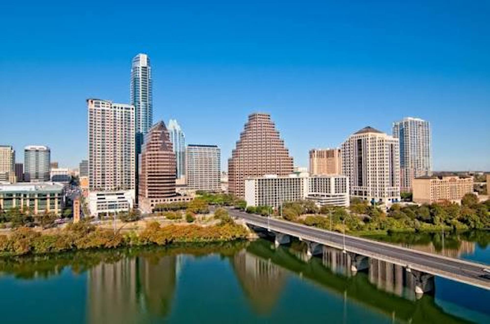 Austin, Texas skyline with modern high-rises, a bridge crossing the reflective Colorado River, lush trees, and a clear blue sky.